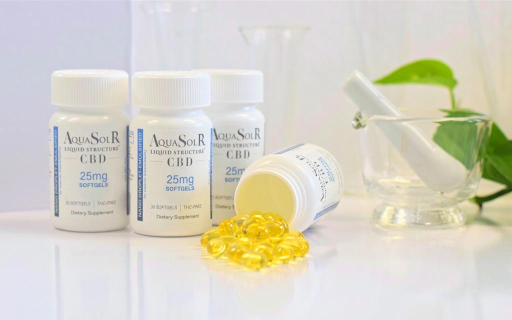 An array of white AquaSolRx CBD bottles with one bottle tipped over and some of the golden-hued liquid-filled gel capsules spilling out of it in a white laboratory setting