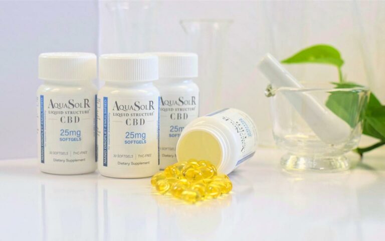 three bottles of AquaSolR CBD softgels sit on a shiny white countertop, with one more bottle tipped over and yellow softgels spilling out of it onto the counter. a mortar and pestle sit to the right side with green leaves from a plant are visible in the background