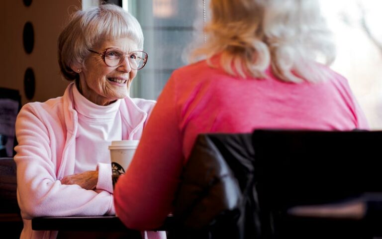 image of two women sitting at a coffee-shop table having a conversation. Closest to us is the back of a woman wearing a pink long-sleeved sweater and her seat back with a black coat draped over it. Across the table from her is an elderly woman with short grey hair in a bob, wearing glasses and smiling, She is wearing a light pink fleece jacket with a matching turtleneck.