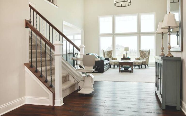 image of a view past a carpeted staircase with hand railings into a light, brightly lit living room with huge floor-to-ceiling windows on the far wall. At the bottom of the central staircase is a stair lift chair in the down position, ready for someone to securely ride up the staircase, or to use the controls at the top of the stairs to bring the stair lift chair to the top and ride it down