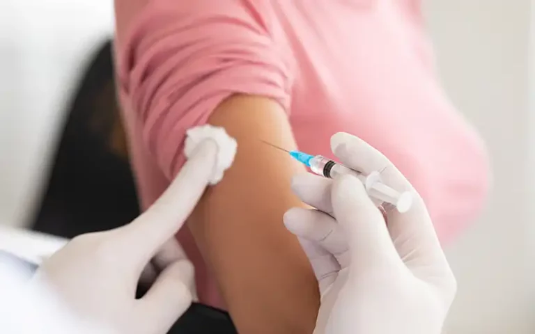 two hands wearing latex gloves are administering a shot in the right arm of a female patient. The patient's pink shirt sleeve is rolled up, and the nurse's left had holds a white cotton ball while the right hand holds a syringe that is ready to inject the vaccine into the patient's arm