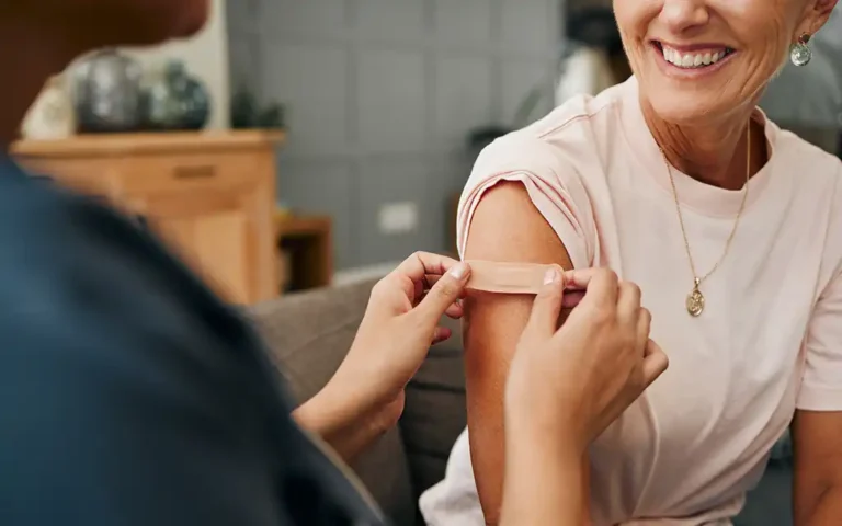 A nurse applies a band-aid to an older woman's arm after she received a vaccination shot. The patient's sleeve is rolled up and we can see the lower half of her face and that she's smiling.