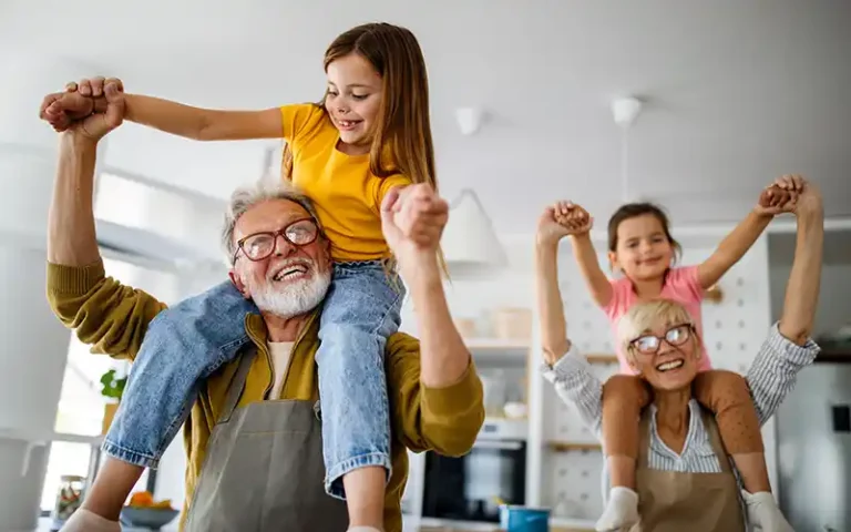 Two people with grey hair and glasses that look like grandparents are in a well-lit light colored kitchen. The Grandfather is on the left and the grandmother is on the right. On each grandparent's shoulders sits a granddaughter, and the grandparent's hands are up, holding hands with the grandchild. All four people are smiling as if they are having a good time.